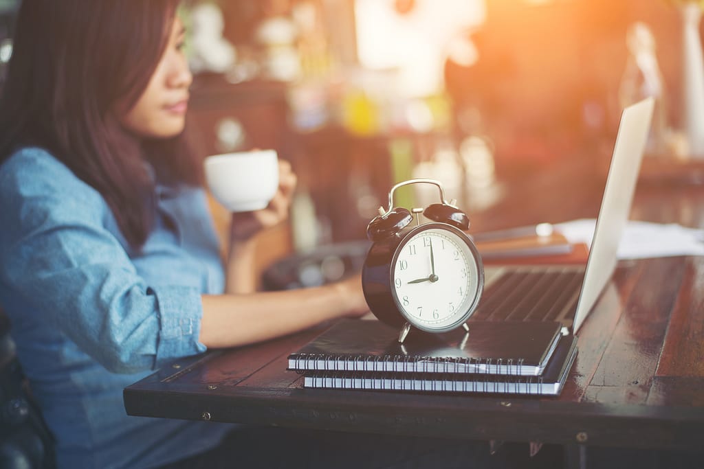 Woman holding coffee while working on a laptop, a clock on the desk symbolizing time reclaimed through a 4-day work week.