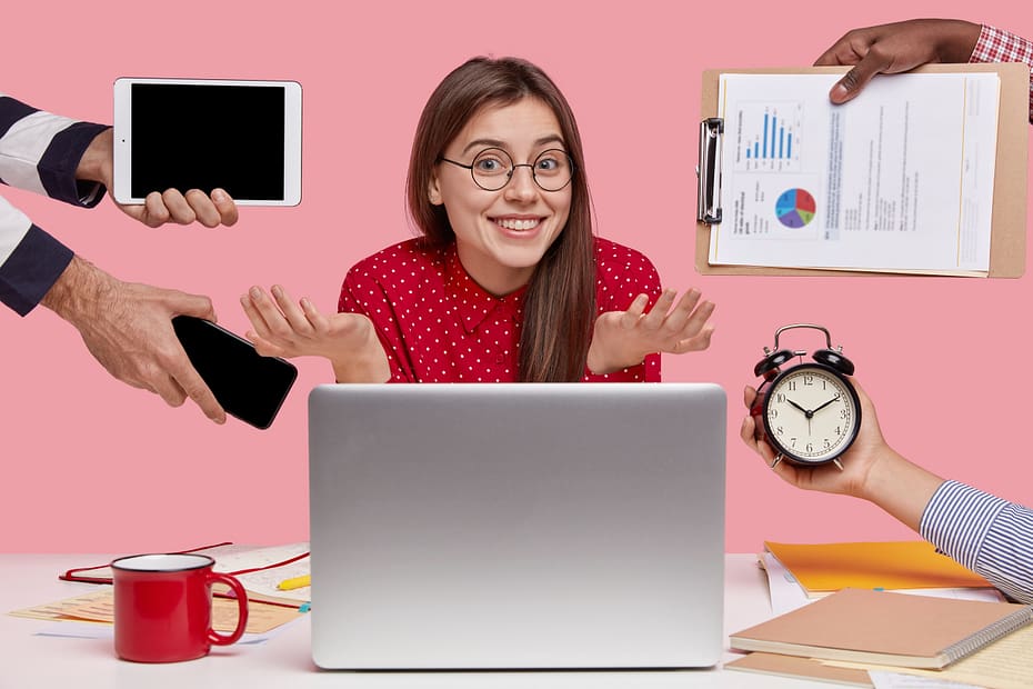 Smiling beautiful dark-haired young woman spreads her palms and smiles broadly with an uncertain facial expression while using a laptop computer to prepare project work, surrounded by modern gadgets and papers, illustrating factors affecting the productivity.