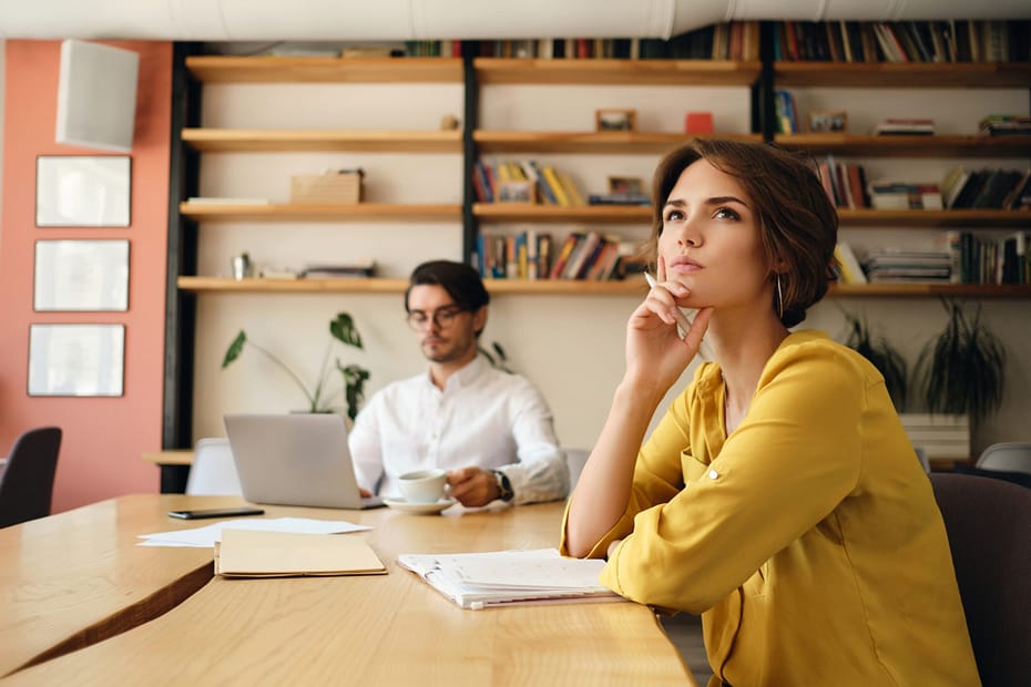Young woman sitting at the table with notepad dreamily looking aside working with colleague on background in modern office